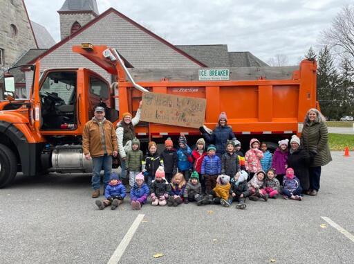 A small group of school children dressed for cold weather sit in front of an orange plow/sander truck. They are holding up a cardboard sign which says, I believe, thank you.