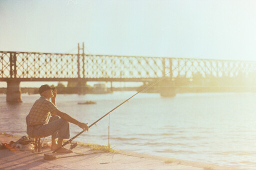 A man is sitting by the riverside, fishing. There is a bridge visible in the background.