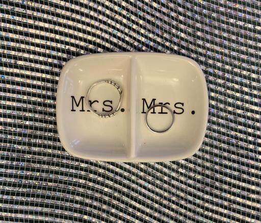 A sparkly tablecloth with a white ceramic ring holder with two silver rings in it, one on each side, one larger than the other. The two sections read Mrs. and Mrs.