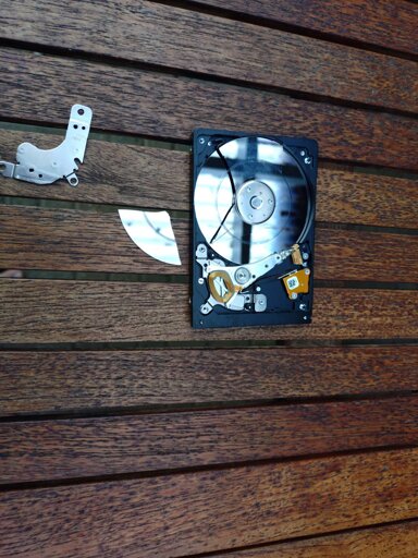 Half a HDD, with the platter exposed. There is visible scrapes from a head crash on the disk, and the top disk in the stack has half of it broken off, sitting next to it. The read/write head is bent up 90°. 
