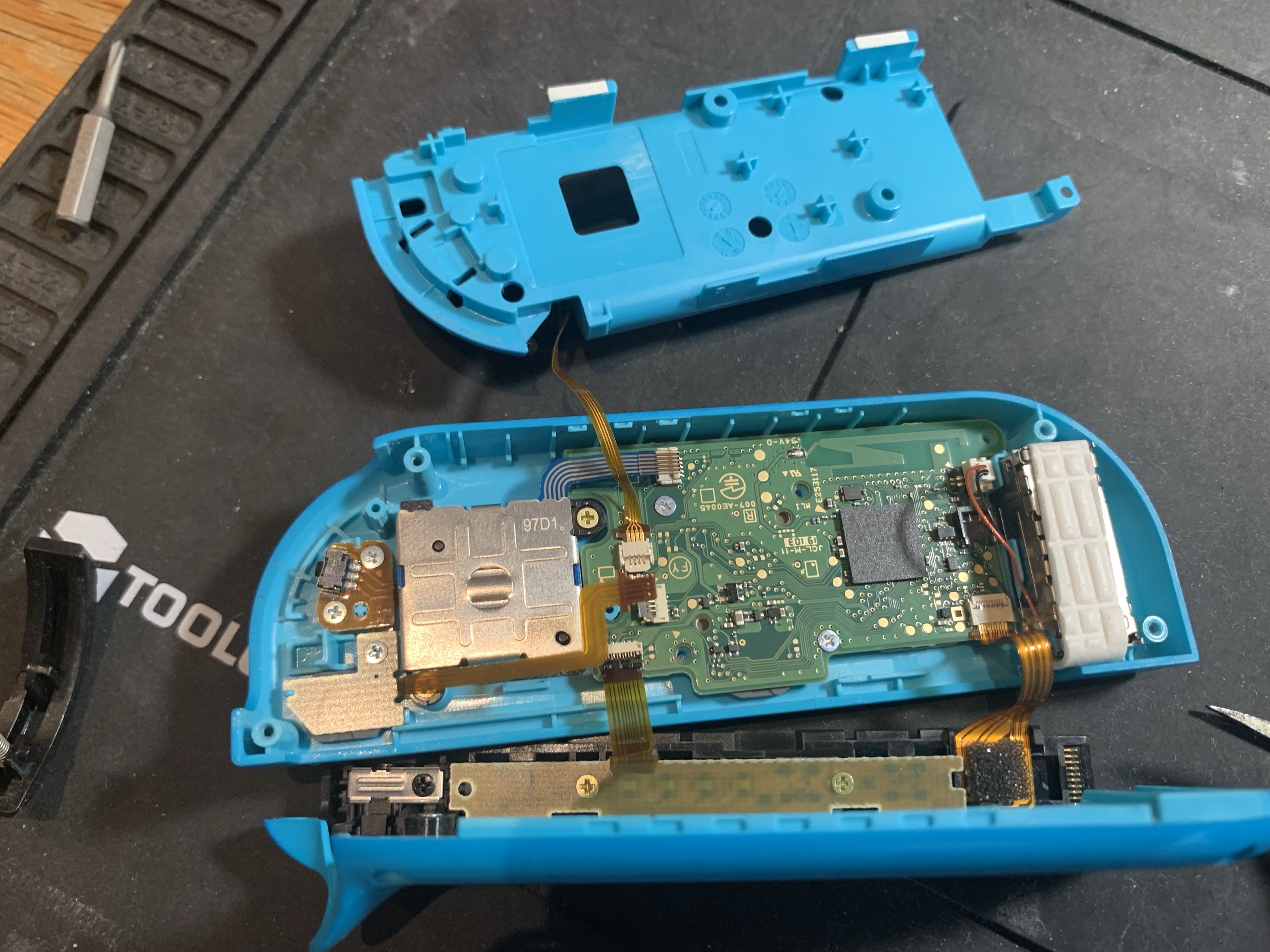 An electronic device is partially disassembled, featuring a blue case, a circuit board with various components, and a metallic cover. Tools are visible in the background on a textured surface.