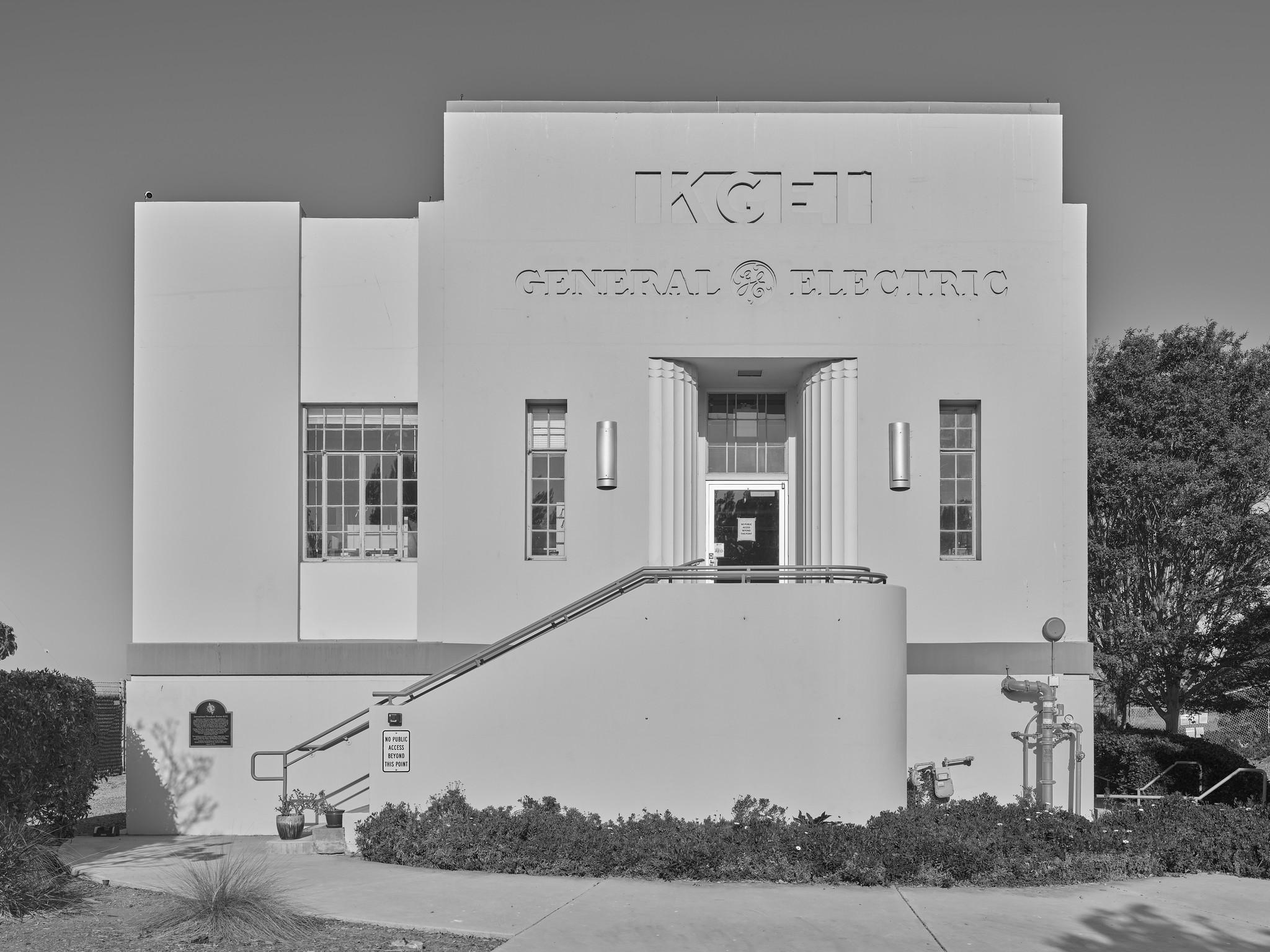 A small concrete building with a staircase leading to a door set back by columns. Above the door, "KGEI / General Electric" is inlayed into the concrete.