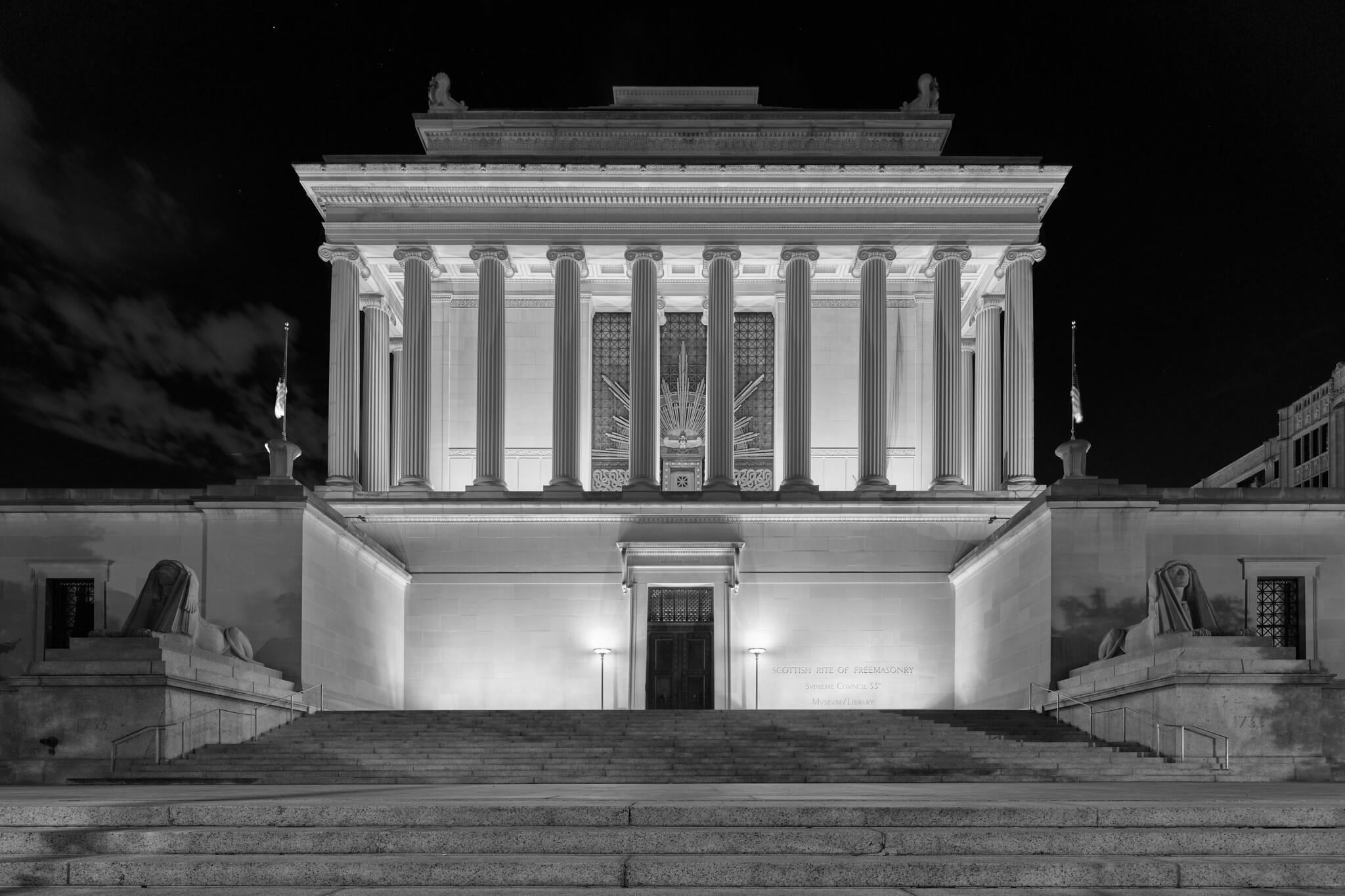 A large Masonic temple at night, with Greek columns, ornate features, and flanked by lions.