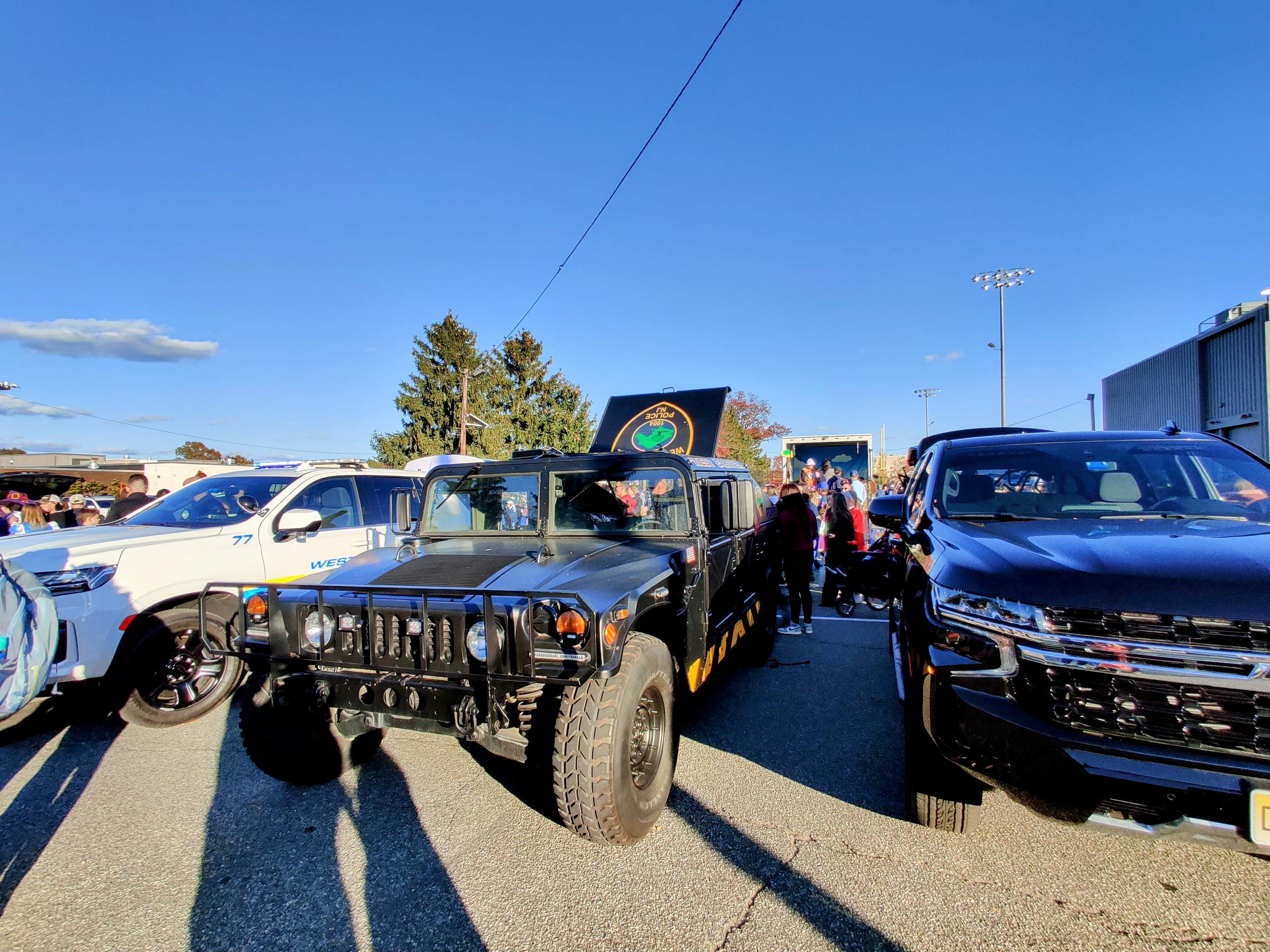 A surplus HUMVEE in black and yellow police livery parked between two new Chevrolet Blazers at a trunk-or-treat event.