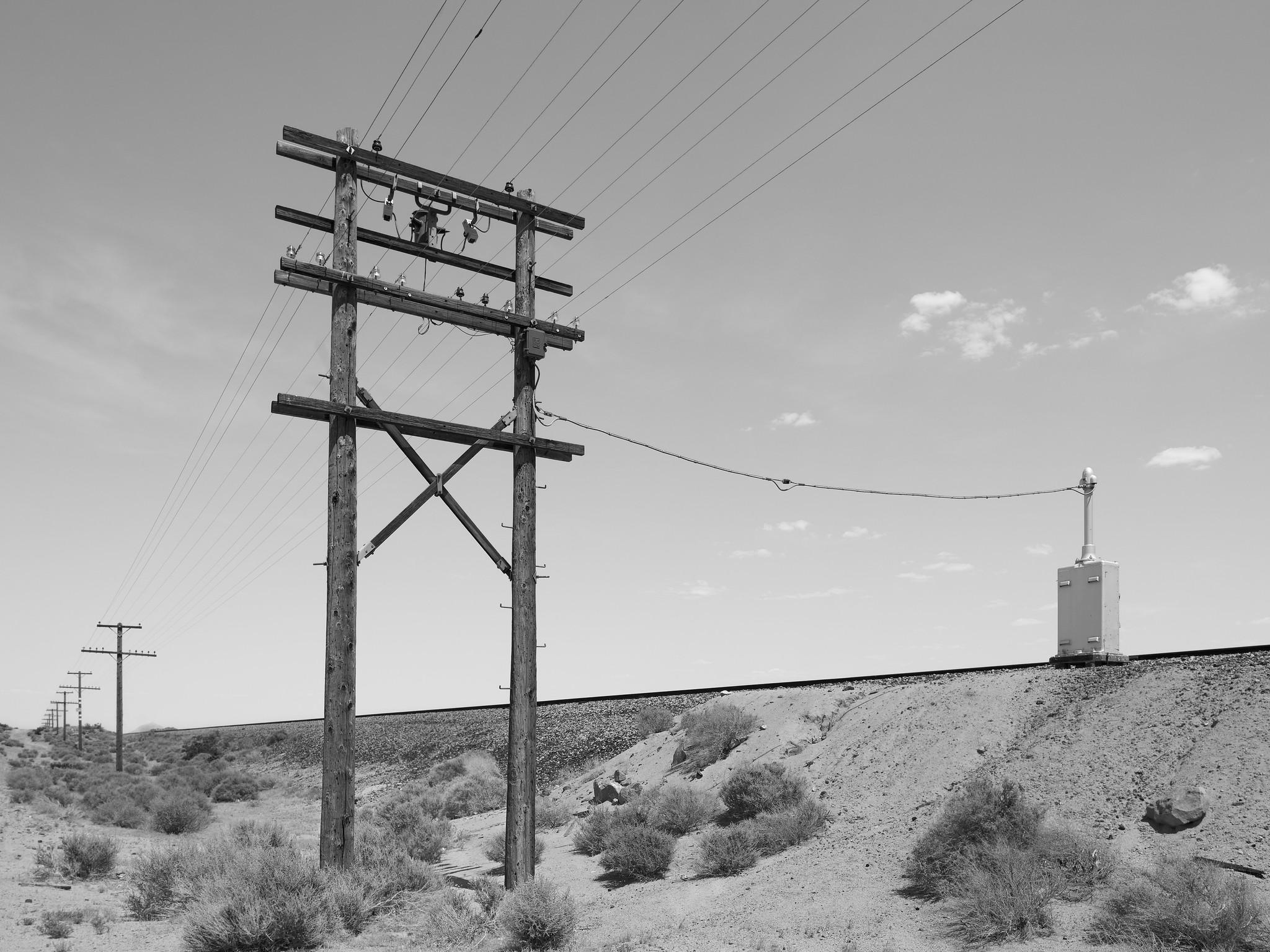 Wooden pylons supporting multiple telegraph and power lines, running adjacent to a railroad track in a barren desert.