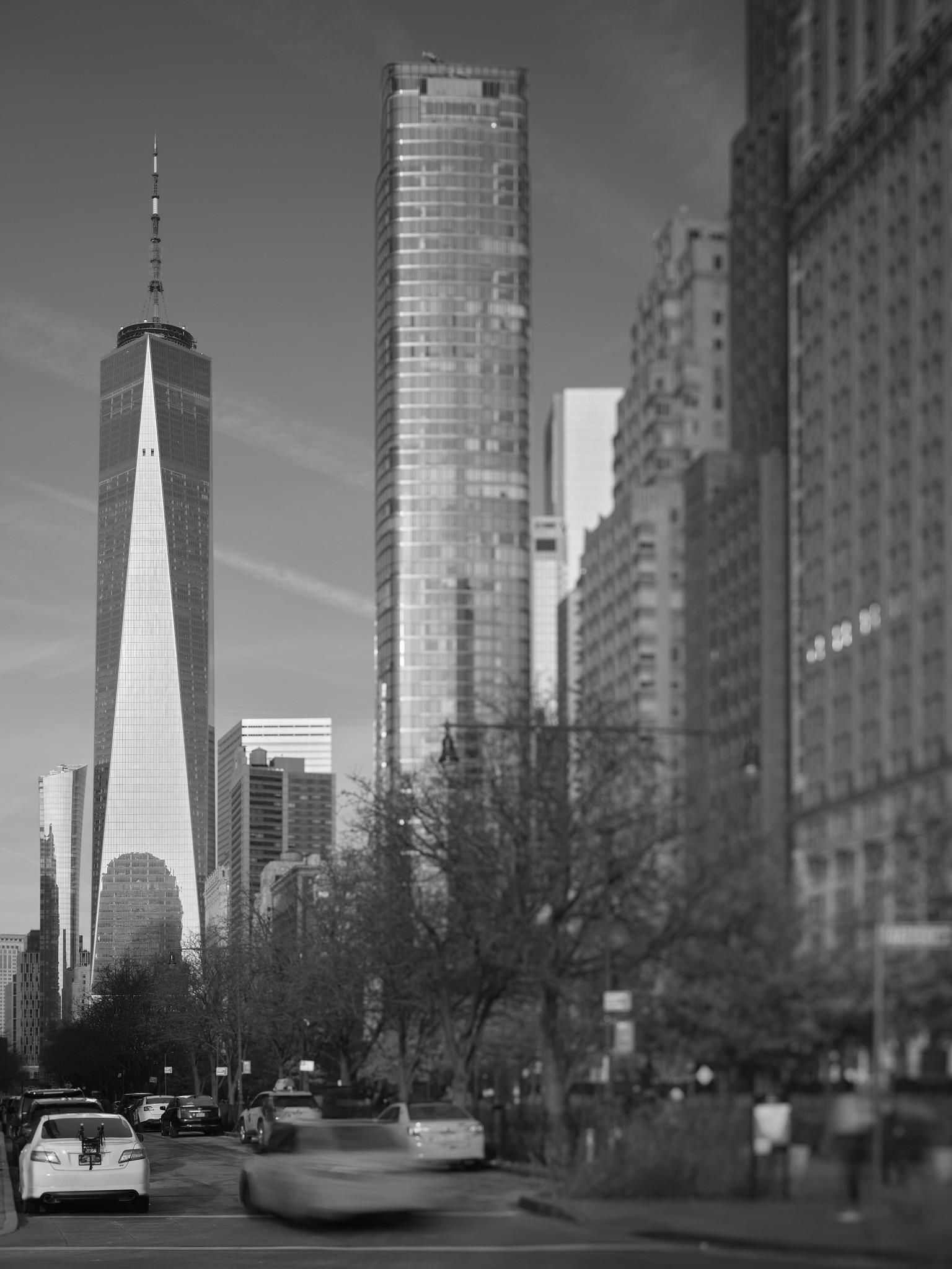 A distinctively shaped reflective skyscraper with a prominent broadcast mast on top, set apart in against the sky at left. Other buildings, in softer focus, crowd the frame at right.