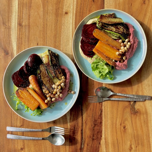 Two light blue glazed plates on a wooden table, garnished with sliced beetroot, grilled carrots and slices of squash, on a bed of light brown and pink hummus, salad slices and grilled chickpeas