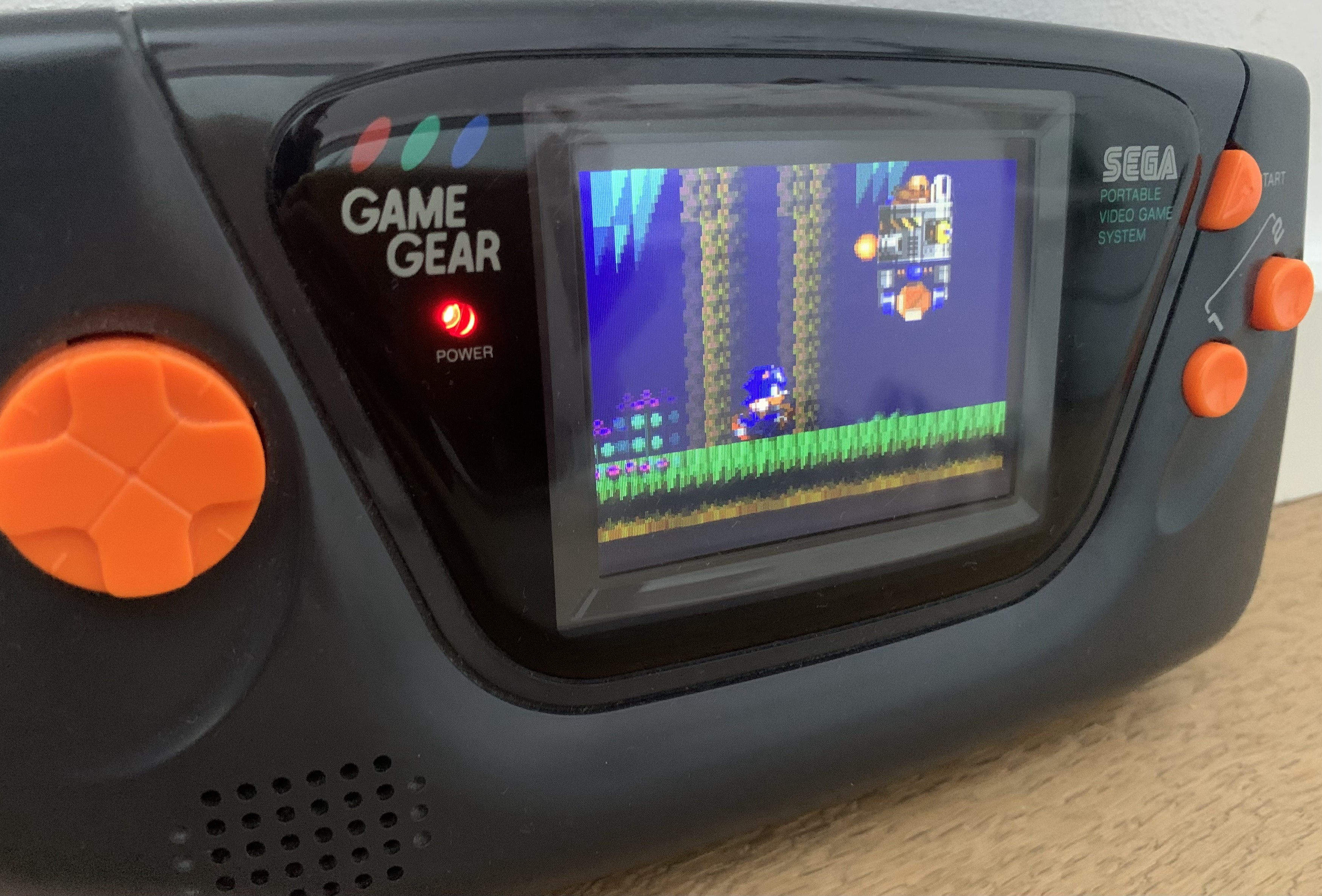 A black Sega GameGear handheld console with black case, bright orange buttons. It’s displaying the opening sequence of Sonic Chaos. Sonic is chasing Eggman who is escaping in a jet powered vehicle carrying a chaos emerald. 